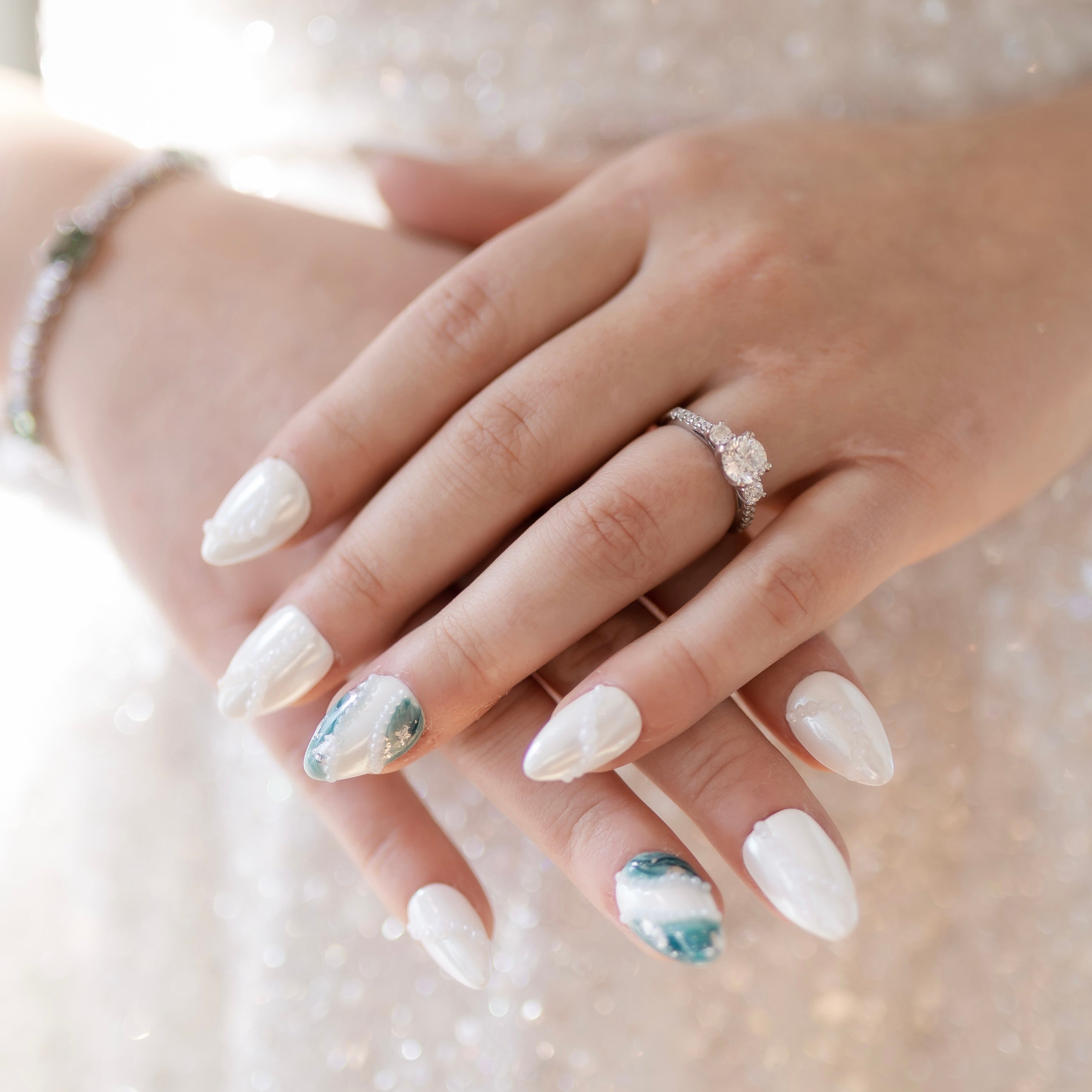 Caucasian woman's hands with press-on nails. The nails are white with a soft pearlescent finish, featuring 3-D pearls arranged in flowing lines. The ring finger has a green marble design with silver foil accents. The marble is in 2 spots: along the left side of the nail, going from the side of the nail right before the cuticle to the tip of the nail, and on the left side starting halfway along the cuticle line and flowing halfway up the nail, ending where the free edge of the nail begins. 